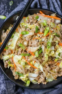 Colorful egg roll in a bowl with ground meat, shredded cabbage, and carrots, garnished with green onions and sesame seeds